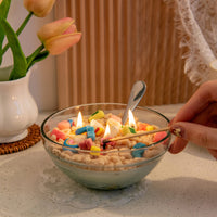 A person lighting the Southlake Gifts Lucky Charms Cereal Candle 2.0, showing bright flames among rainbow and clover wax charms — a whimsical handmade candle perfect for gifting.