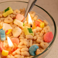 Close-up of the Lucky Charms Cereal Candle 2.0 burning, showing colorful marshmallow wax shapes like rainbows, hearts, and stars — a detailed novelty candle that makes a magical artisan gift.