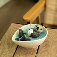 Southlake Gifts Floating Otter Candle lit and glowing softly indoors, showing the otter holding a seashell with warm candlelight reflection, an artisan gift for ocean and animal lovers.