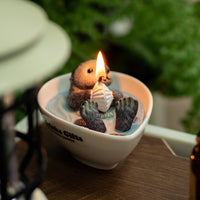Close-up of the Southlake Gifts Sea Otter Candle surrounded by green plants, showing a realistic otter holding a seashell in blue wax, a cozy artisan décor piece and thoughtful gift.