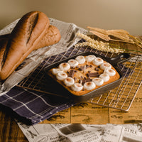 Southlake Gifts Skillet S’mores Candle styled on a rustic baking scene with bread, cooling rack, and newspaper backdrop, a realistic chocolate s'more marshmallow dessert candle in a real skillet pan with cookie base, designed as a unique novelty candle and perfect foodie gift for birthdays, housewarmings, and food s'more lovers.