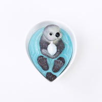 Top-view of the Southlake Gifts Sea Otter Candle in a white ceramic bowl, featuring an otter holding a seashell surrounded by blue wax waves, a realistic handmade candle ideal for gifting during holidays or housewarmings.