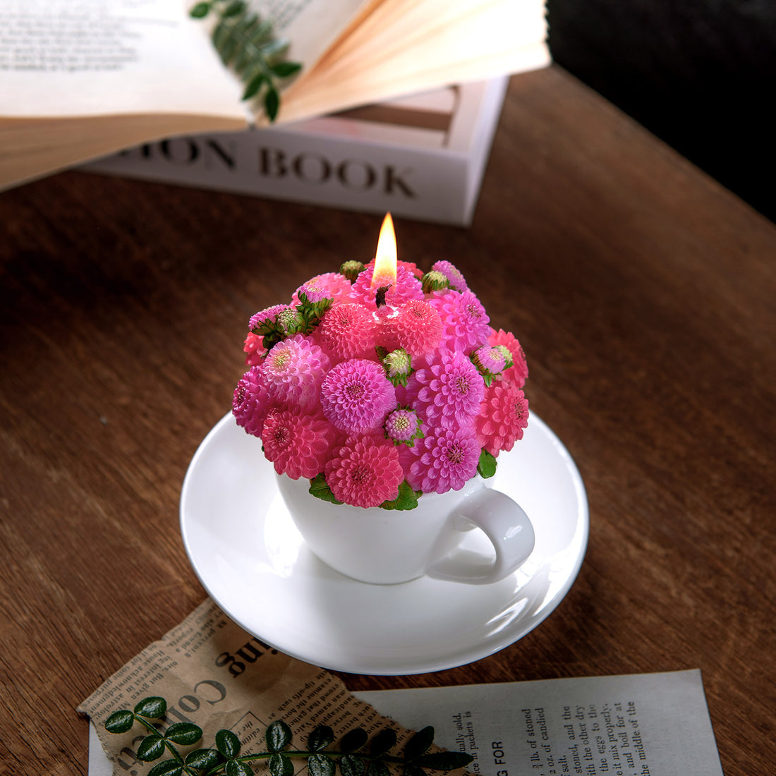 Southlake gifts Dahlia floral aromatherapy candle designed to look like a bouquet lit up, set on a white cup and saucer on a wooden surface with a book in the background.
