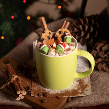 Southlake Gifts Fireside Cocoa Candle Mug lit on a cozy table with pinecones and cinnamon sticks, featuring realistic soy wax chocolate chip cookies, fluffy marshmallows, cinnamon stick wicks, and green marshmallow Santa hat wax melts — handmade holiday candle décor.