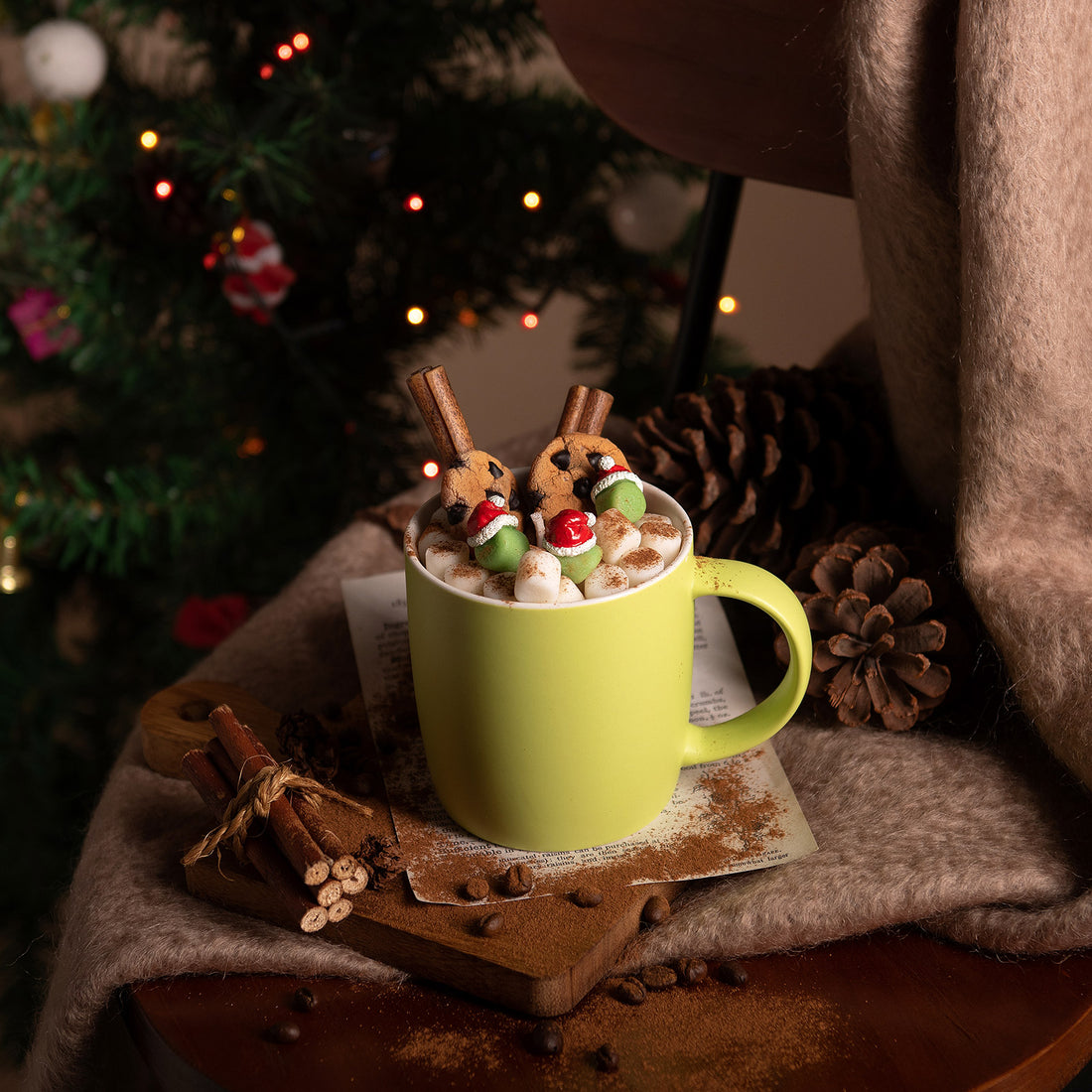 Festive hot cocoa candle mug with flickering flame surrounded by Christmas lights — handcrafted soy wax cookies, cinnamon sticks, and marshmallow melts in a reusable green ceramic cup.