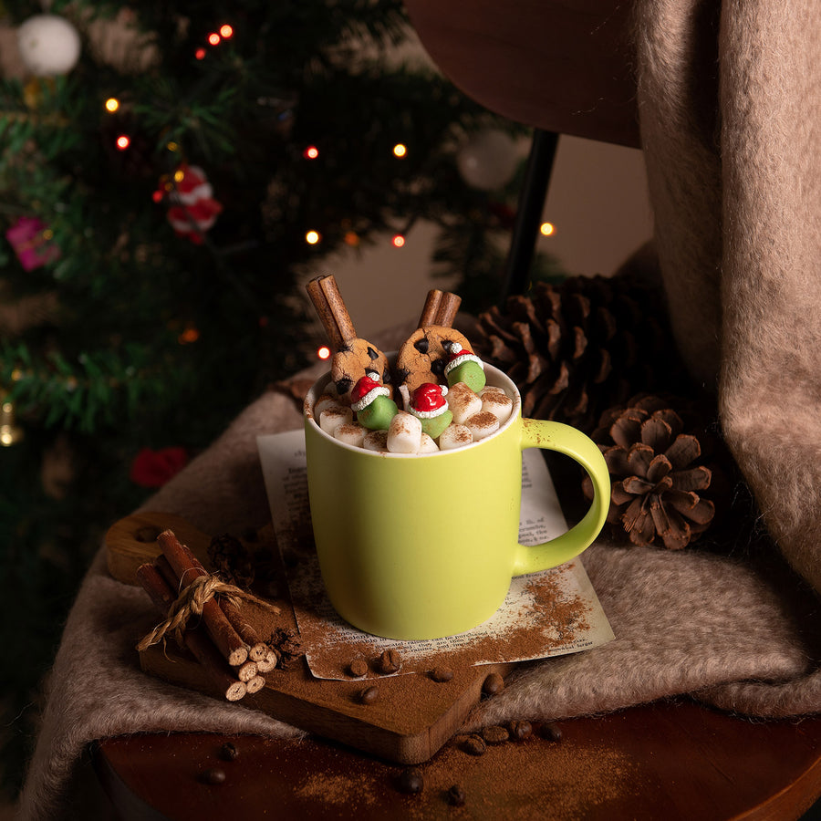 Festive hot cocoa candle mug with flickering flame surrounded by Christmas lights — handcrafted soy wax cookies, cinnamon sticks, and marshmallow melts in a reusable green ceramic cup.