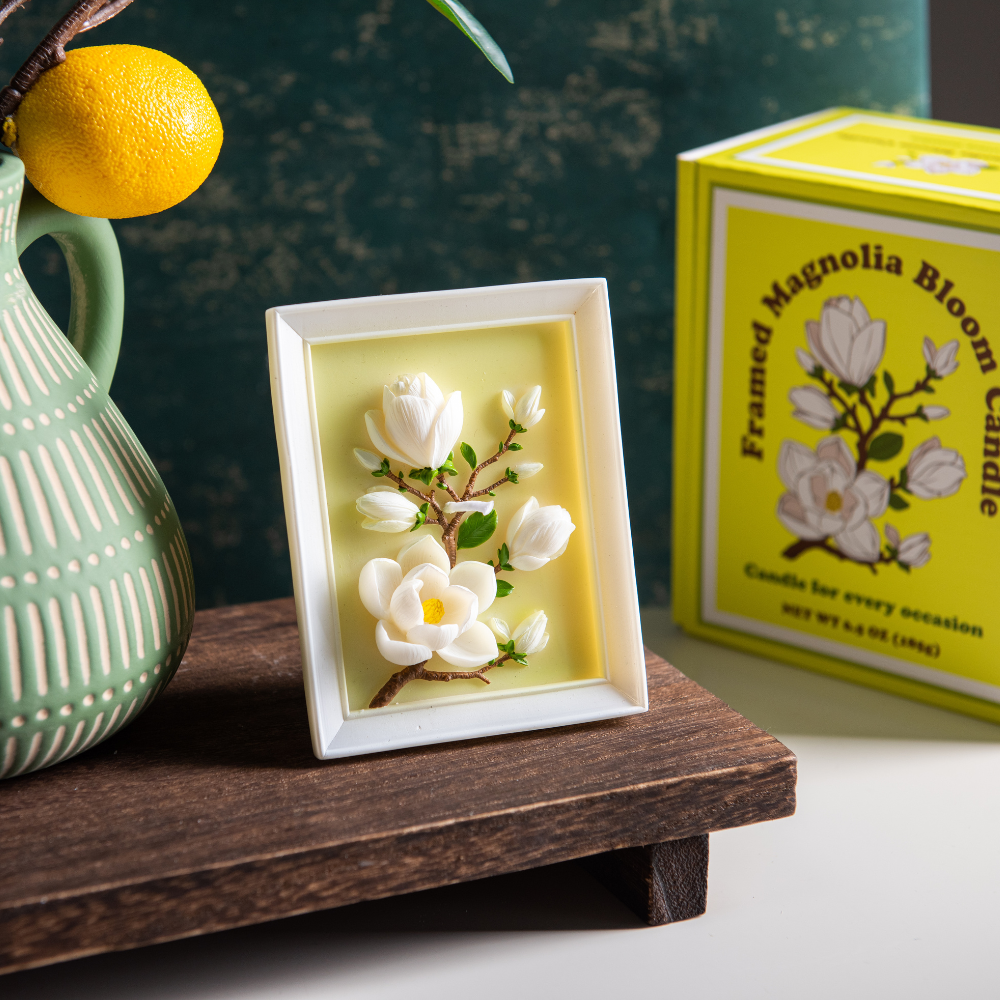 Southlake Gifts Framed Magnolia Bloom Candle displayed upright as a botanical frame candle with its gift-ready packaging on the side, featuring hand-sculpted magnolia flowers in a white picture-frame design, an elegant handmade soy wax candle for refined home décor and a thoughtful gift for any season.