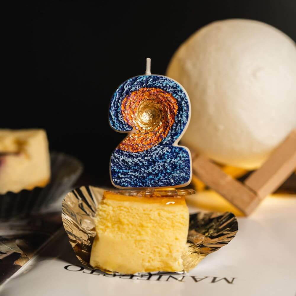 Southlake Gifts Galaxy black hole number 2 candle displayed on cheesecake slice with warm moon lamp lighting, a space themed birthday cake topper and novelty number candle.