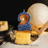 Southlake Gifts Galaxy black hole number 2 candle displayed on cheesecake slice with warm moon lamp lighting, a space themed birthday cake topper and novelty number candle.