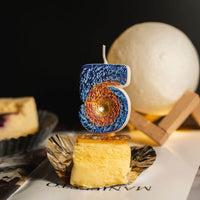 Southlake Gifts Galaxy black hole number 5 candle displayed on cheesecake dessert with moon lamp backdrop, a space themed birthday cake topper and novelty candle.