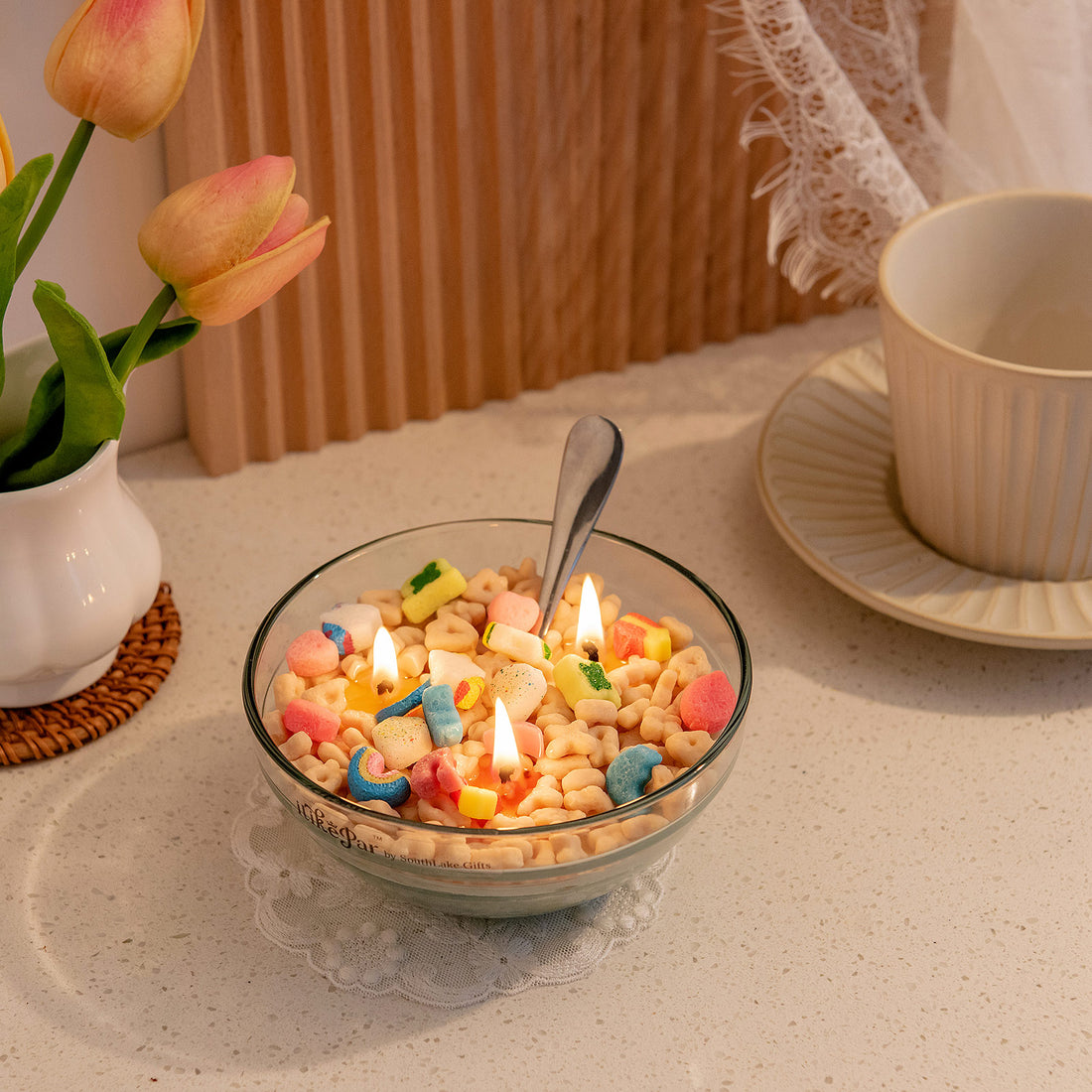 Southlake Gifts Lucky Charms cereal bowl candle with multicoloured charms resembling Lucky Charms cereal pieces, lit and placed on a table with a silver metal spoon right inside it.