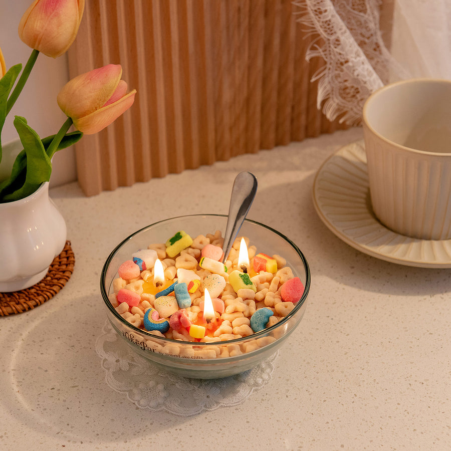 Southlake Gifts Lucky Charms cereal bowl candle with multicoloured charms resembling Lucky Charms cereal pieces, lit and placed on a table with a silver metal spoon right inside it.
