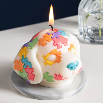 Southlake Gifts Puppy Puzzle Cute Animal Candle is lit up – handmade dog-shaped candle with colorful puzzle pieces, pine scented novelty décor on a white desk 