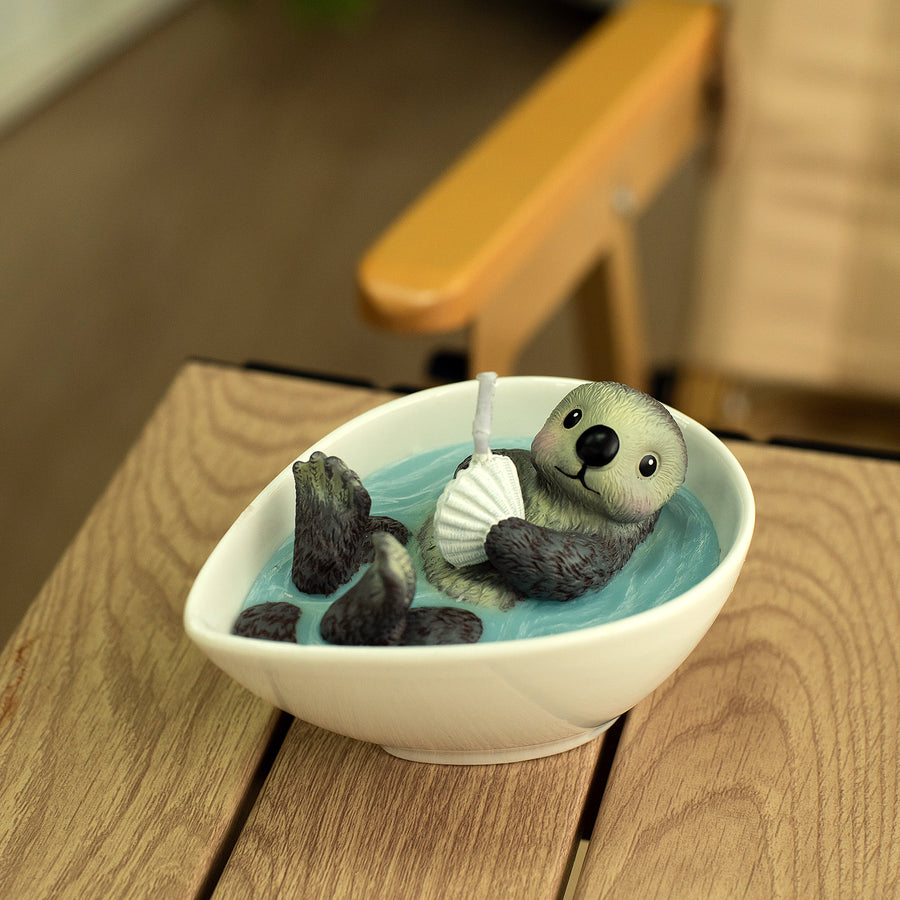 Southlake Gifts Floating Otter Candle lit and glowing softly indoors, showing the otter holding a seashell with warm candlelight reflection, an artisan gift for ocean and animal lovers.