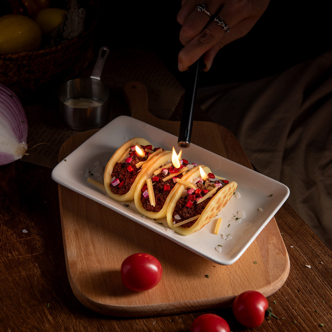 taco candle from Southlake Gifts with a lit flame on a wooden plate, surrounded by ingredients such as tomatoes and onions, and a person's hand is visible holding a spoon in the background.