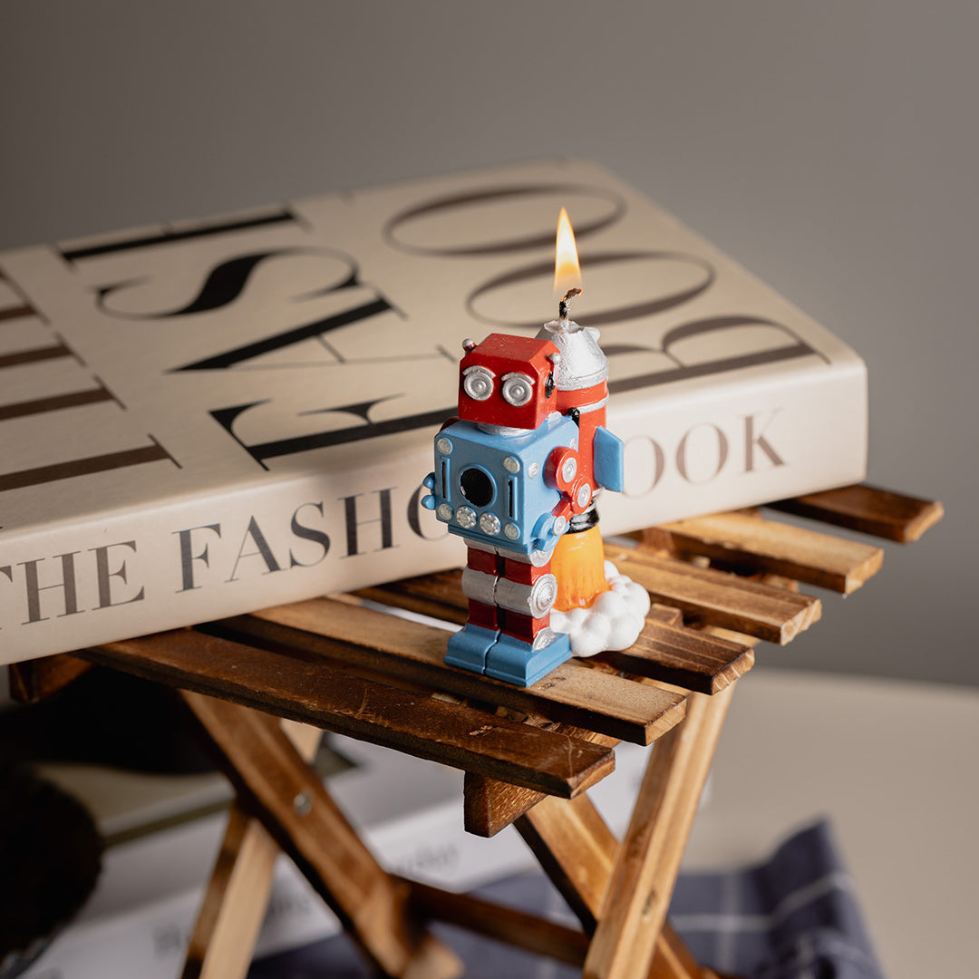 Vintage Robot Rocket Candle Cake Topper   is lit up and stanidng on a desk in front of a book as cute novelty cake/home decoration candle . 