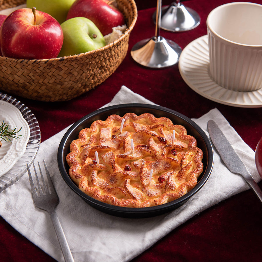 Southlake Gifts Apple pie food candle on a black baking pan with a basket of apples and a cup in the background.