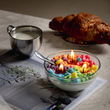 Southlake Gifts Fruit Loops Cereal Bowl Scented food Soy wax Candle with a real spoon is lit up, sitting on a table, a novelty candle decor, unique candle gifts, while a cup of milk and a croissant sit in the background.  