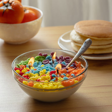 Southlake Gifts Fruity Pebbles Cereal Candle Bowl with metal spoon is lit and sitting on a wooden table with pancakes in the background — a realistic handmade food candle that makes a playful breakfast-inspired gift.