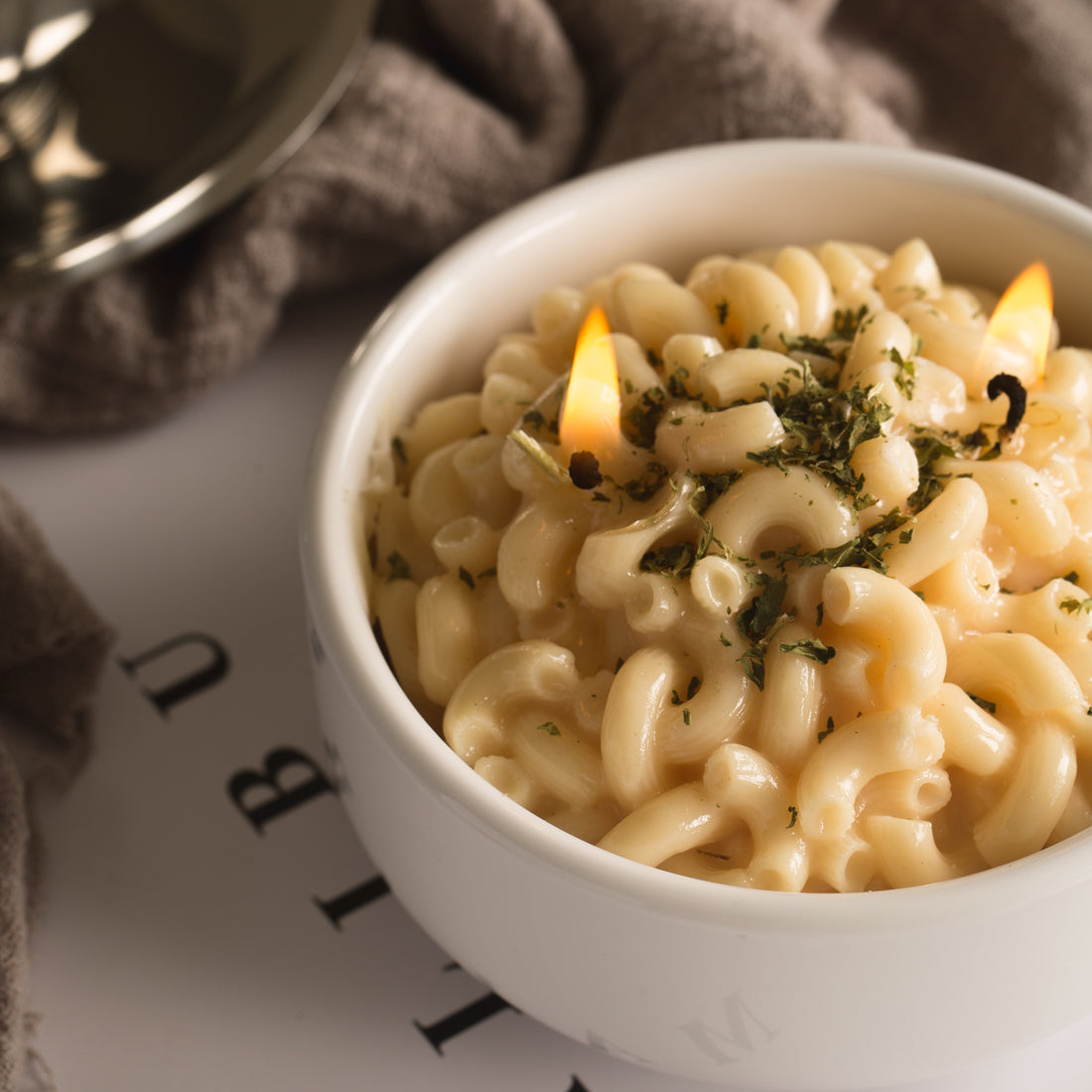 Close-up of Southlake Gifts Macaroni & Cheese Pasta Candle lit with two flames — realistic soy wax scented rigatoni mac and cheese topped with parsley, a handmade candle that makes a warm and playful gift for foodies and candle lovers.