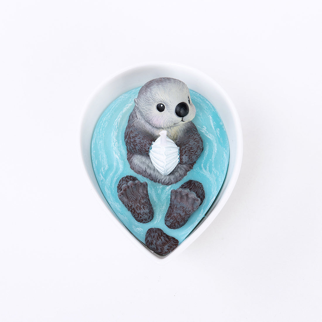 Top-view of the Southlake Gifts Sea Otter Candle in a white ceramic bowl, featuring an otter holding a seashell surrounded by blue wax waves, a realistic handmade candle ideal for gifting during holidays or housewarmings.