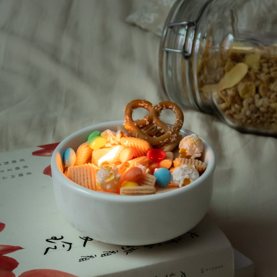 Whimsical snack mix candle lit on a book stack, showing glowing wax pretzel and colorful M&M-style details — a unique handmade candle perfect for holiday gifting.